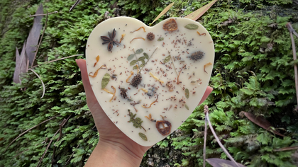 Hand holding Mary Heartful Wax Melt in heart shape with natural botanical decorations by MeaQuasar, showcasing sustainable design with pine, cedar, and patchouli. Eco-friendly wax melt against a green mossy background.